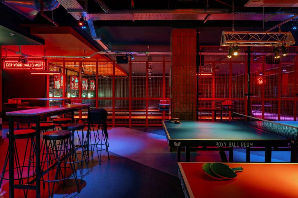 Gaming hall with pool tables, ping pong, bar seating, and red neon lighting throughout industrial space.