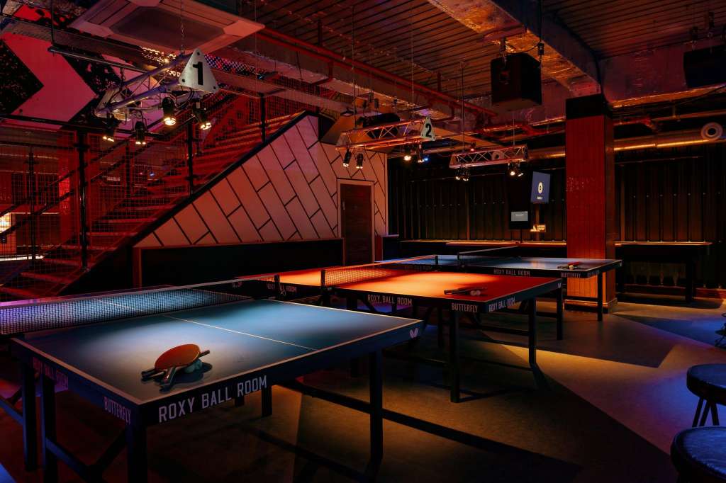 Crazy Pool room with multiple ping pong tables, red and blue ambient lighting, industrial ceiling with exposed beams.