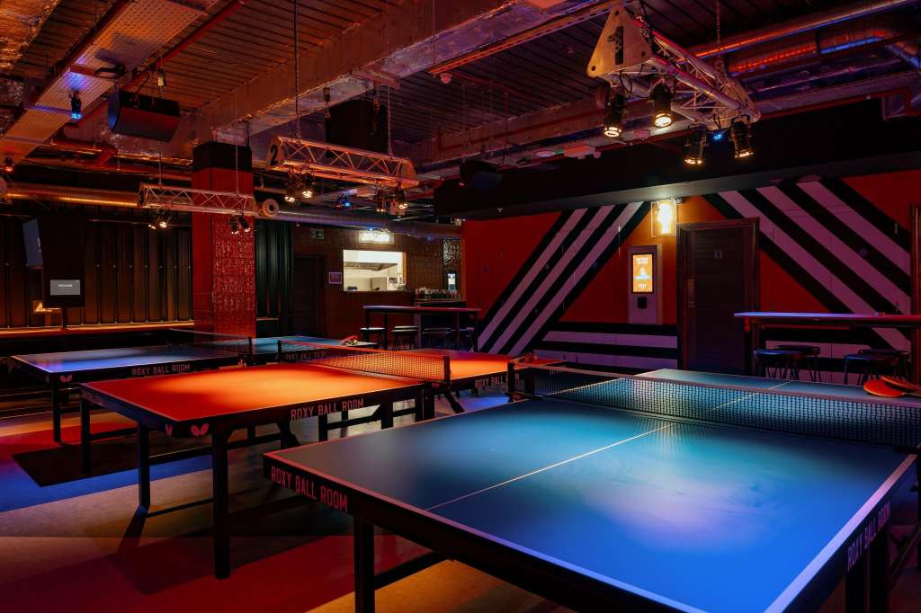 Gaming hall with multiple pool tables lit in blue and orange, industrial ceiling with exposed beams and neon accents.