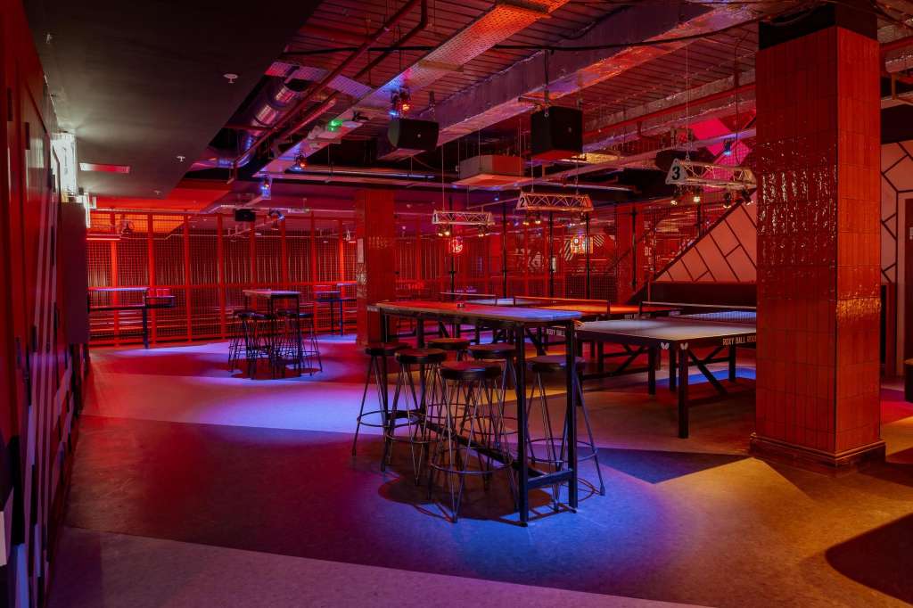Gaming hall with pool tables, high-top seating, and dynamic red and blue ambient lighting throughout.