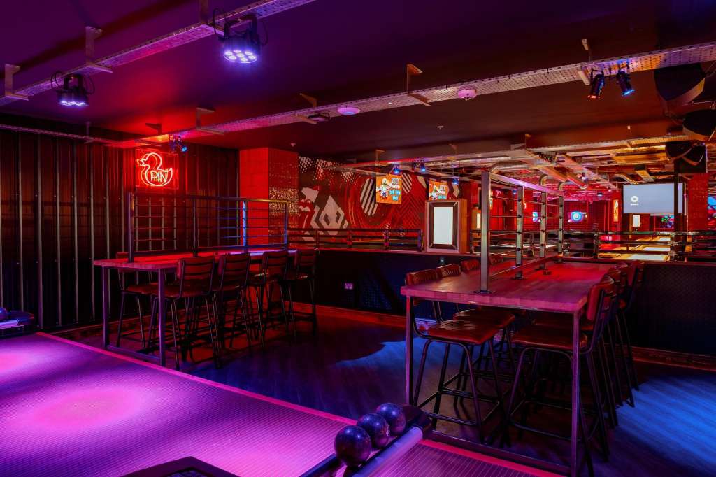 Crazy Pool room with neon lighting, high-top tables, bar seating, and bowling balls on illuminated floor.