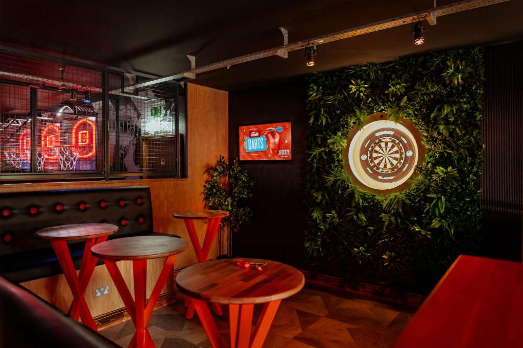 Gaming hall with dartboard, neon signage, red tables, and green plant wall backdrop.