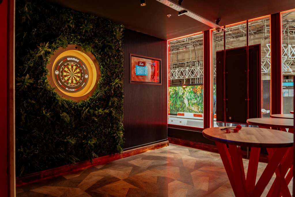 Gaming hall with illuminated dartboard on foliage-backdrop wall, red gaming table, and neon signage.