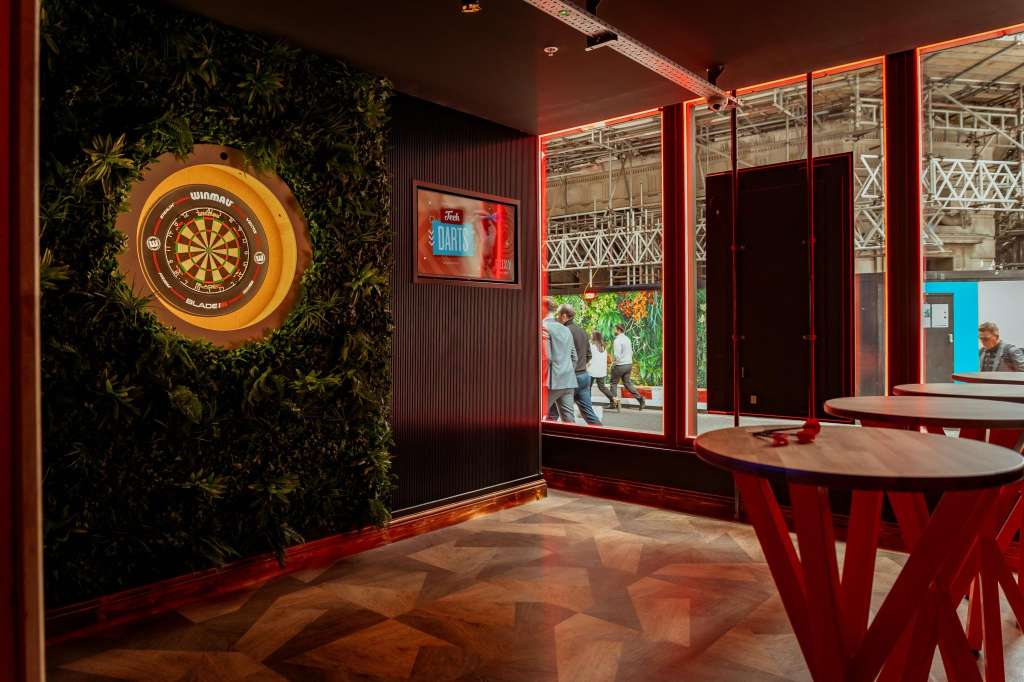 Gaming hall with illuminated dartboard on green wall, red furniture, and geometric flooring.