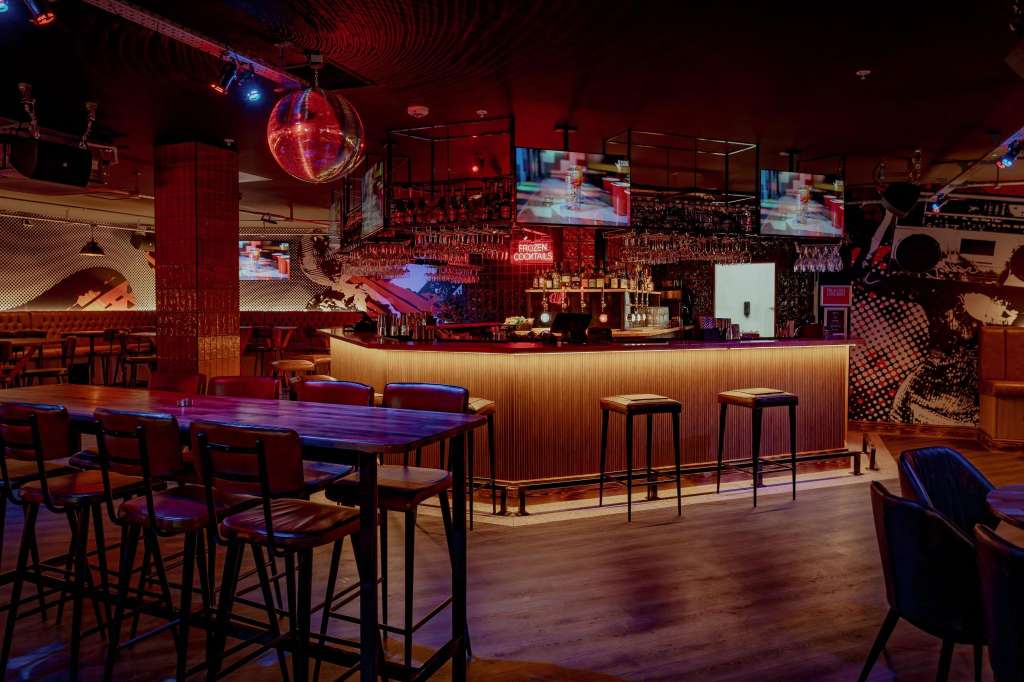 Modern bar and lounge with illuminated counter, high-top tables, red ambient lighting, disco ball, and screens.