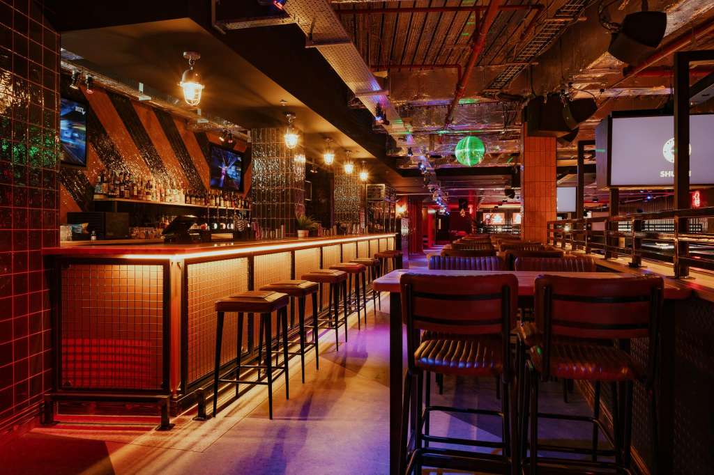 Modern bar with red tile walls, illuminated counter seating, ambient purple and gold lighting, and lounge seating area.