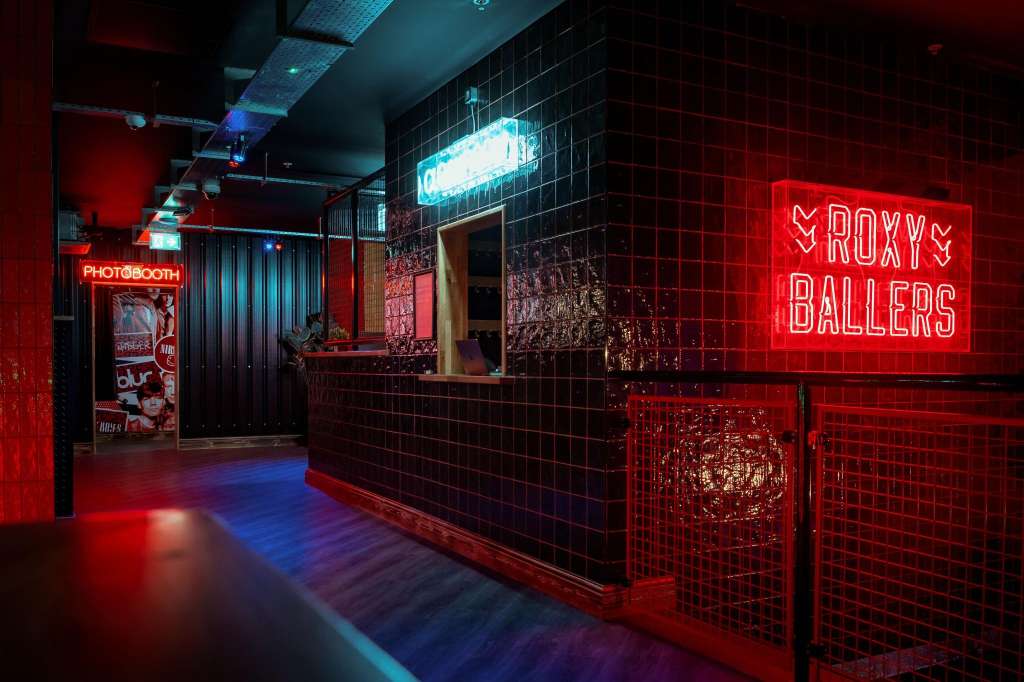 Modern bar area with neon signage, black tile walls, red and blue ambient lighting, and service counter.