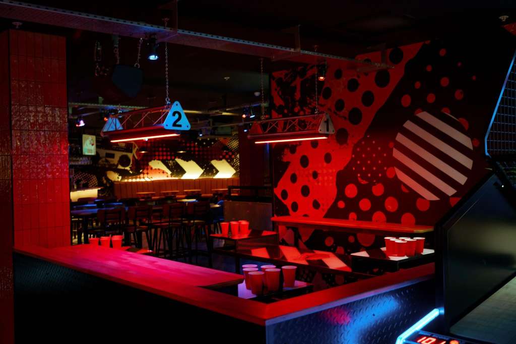 Gaming hall with red and black polka-dot decor, neon lighting, bar seating, and gaming tables.