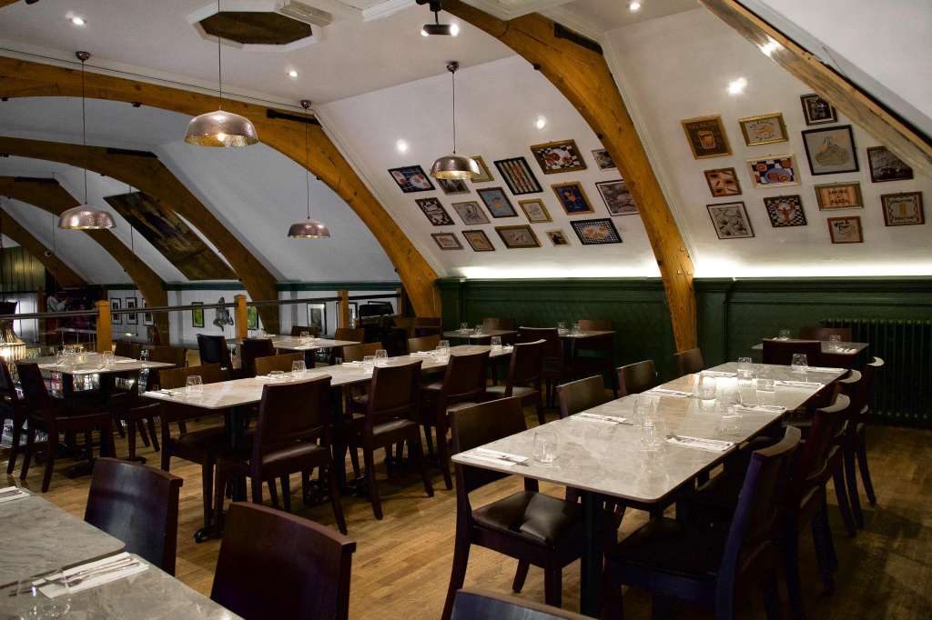 Upscale dining room with exposed wooden beams, pendant lighting, framed artwork gallery wall, and long tables set with place settings.