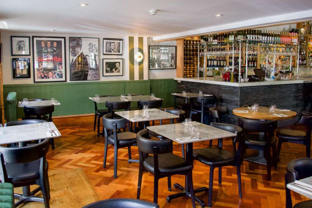 Restaurant dining room with scattered tables, black chairs, green banquette seating, full bar, and framed artwork on walls.