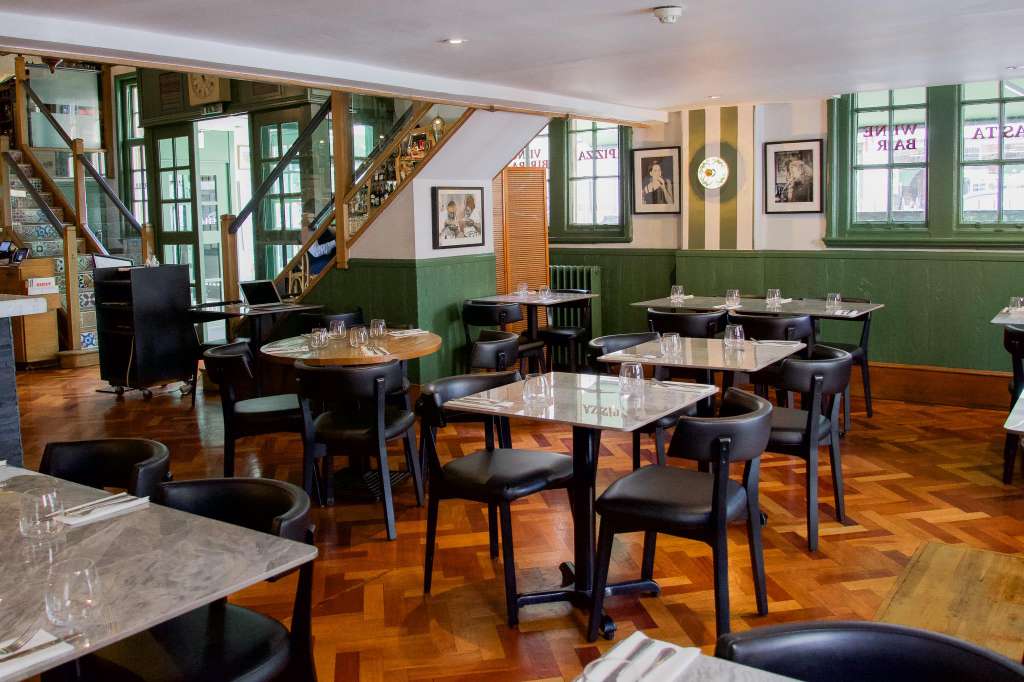Restaurant dining room with multiple tables set with glassware, black chairs, green wainscoting, and wooden staircase.
