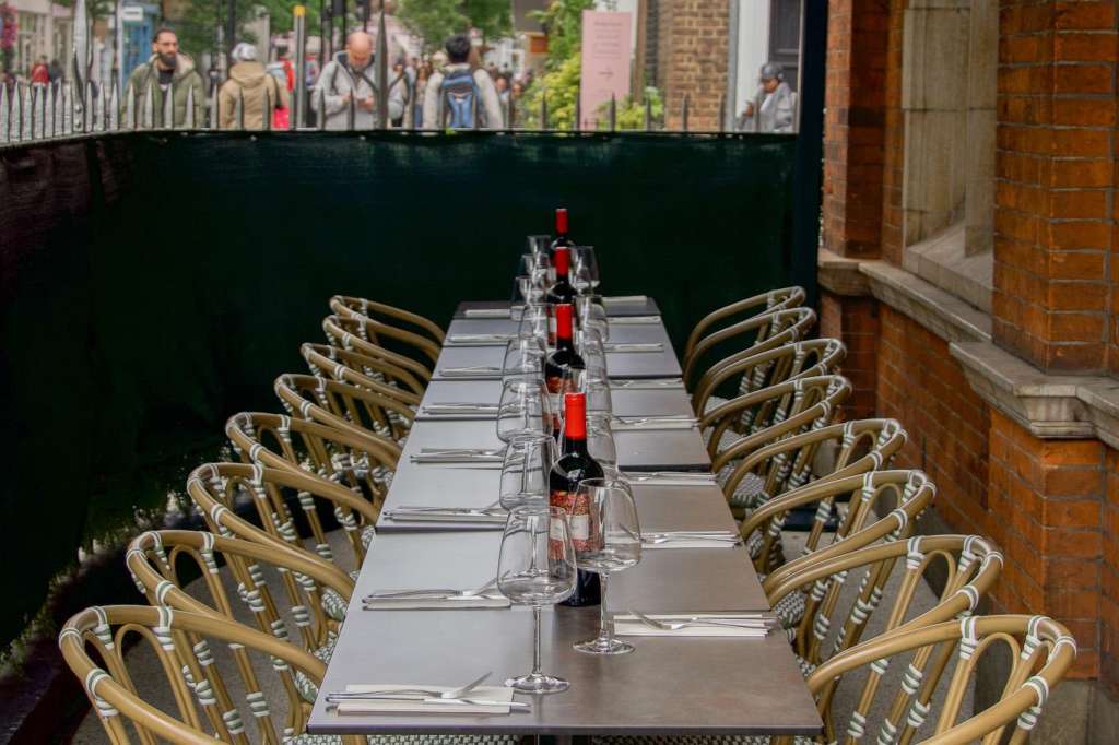 Long outdoor table set for dining with place settings, wine glasses, and rattan chairs under green awning.