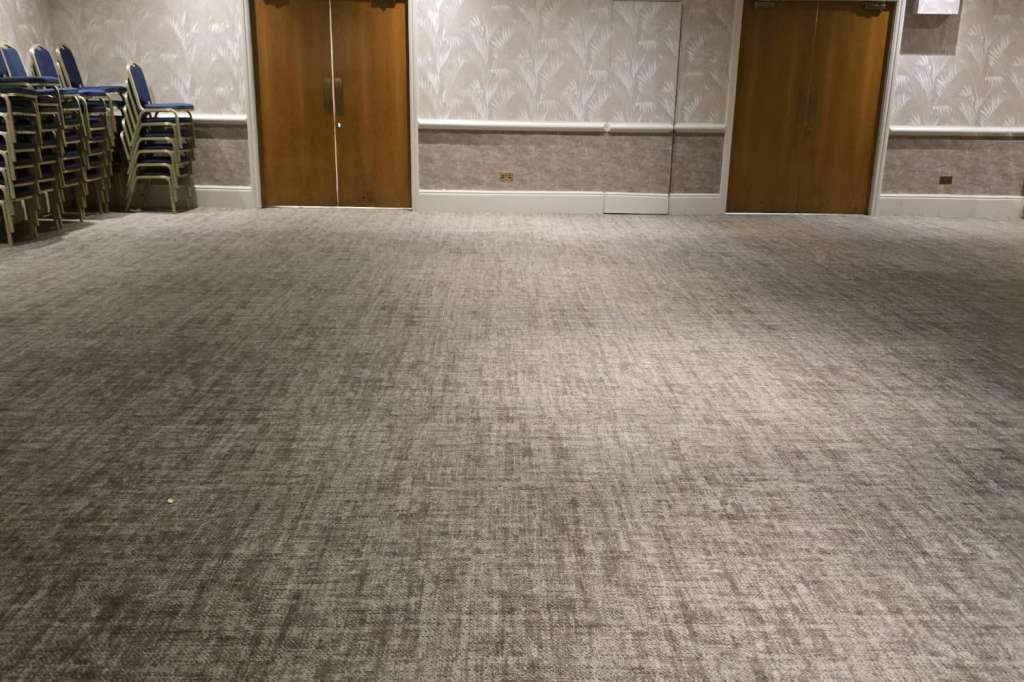 Empty ballroom with gray carpet, decorative chandelier, patterned wallpaper, and stacked chairs along the left wall.