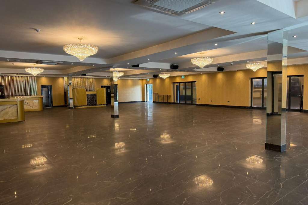 Large empty ballroom with polished dark floor, gold-toned walls, crystal chandeliers, and support columns.