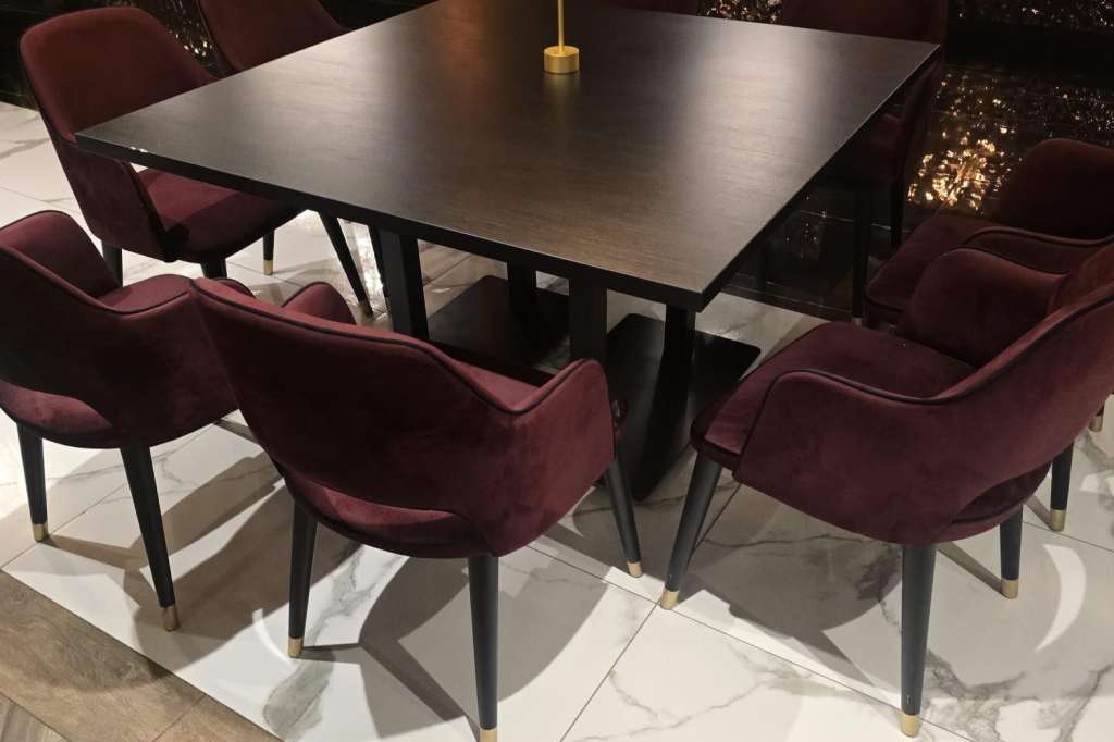Modern dining table with burgundy chairs set in an upscale interior with textured black wall paneling and ambient lighting.