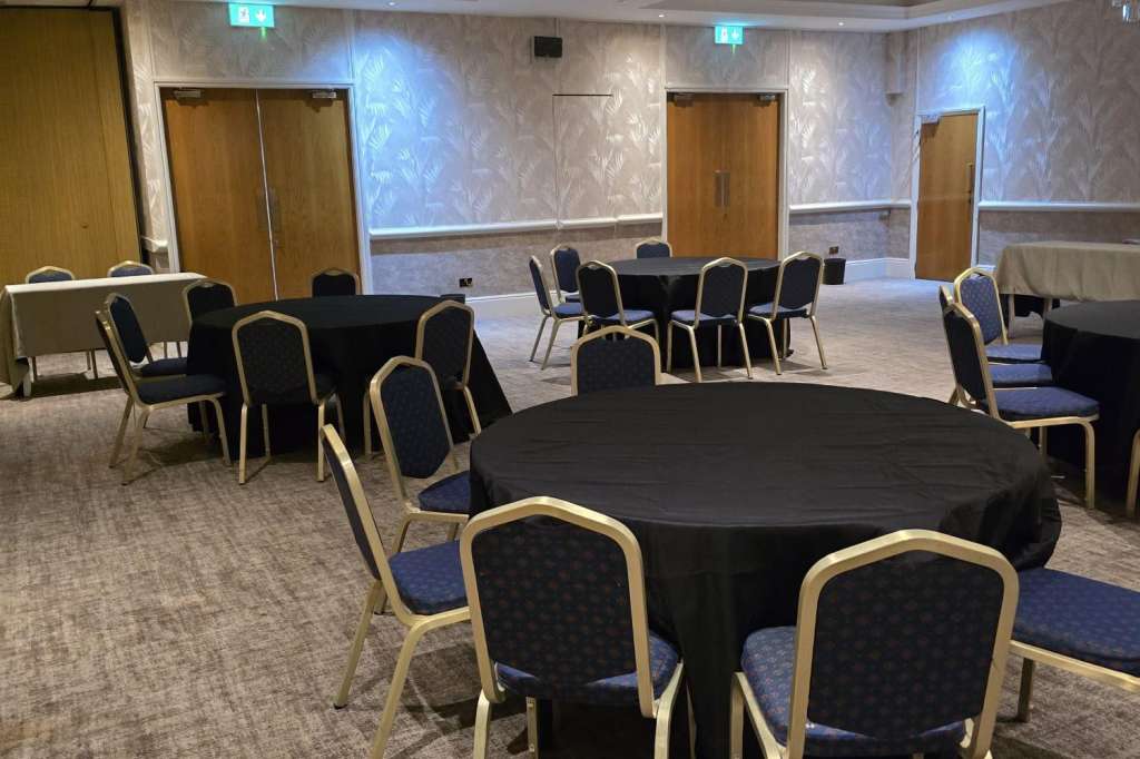Ballroom with round tables dressed in black linens, gold-framed chairs, crystal chandelier, and ambient blue lighting.