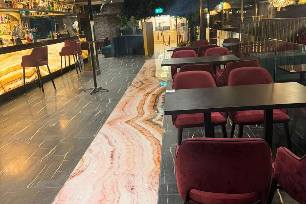 Modern venue space with burgundy chairs at tables, bar area, decorative ceiling, and marble flooring.