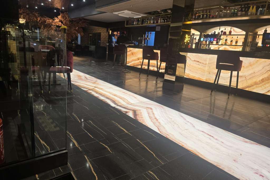 Modern bar and lounge area with illuminated marble counter, black chairs, and backlit bottle display shelving.