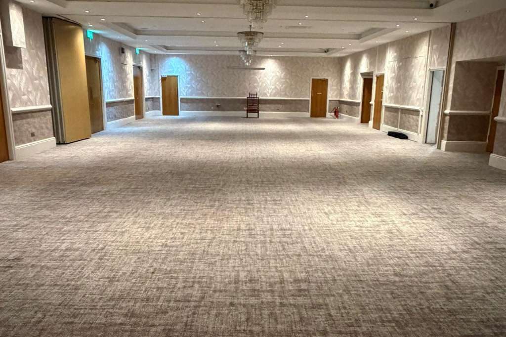 Large empty ballroom with neutral carpet, recessed ceiling lights, crystal chandelier, and multiple doorways.