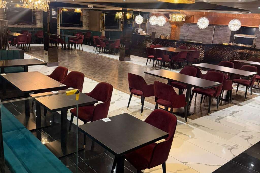Modern dining space with rectangular tables, burgundy chairs, gold pendant lights, and open kitchen visible behind.