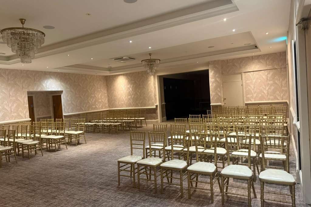 Plaza Suite set up with rows of gold Chiavari chairs with white cushions, crystal chandeliers, and patterned wallpaper.