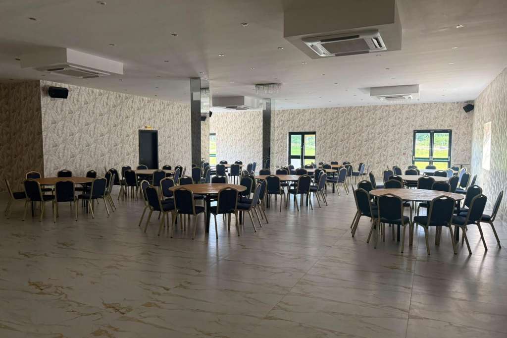 Large open room with round tables, black chairs, and recessed ceiling lighting. Patterned wallpaper and support columns throughout.