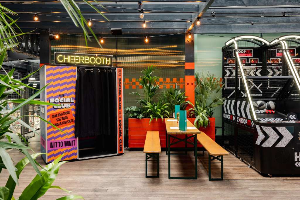 Rooftop venue with cheerbooth, arcade games, plants, and wooden bench seating under warm pendant lighting.