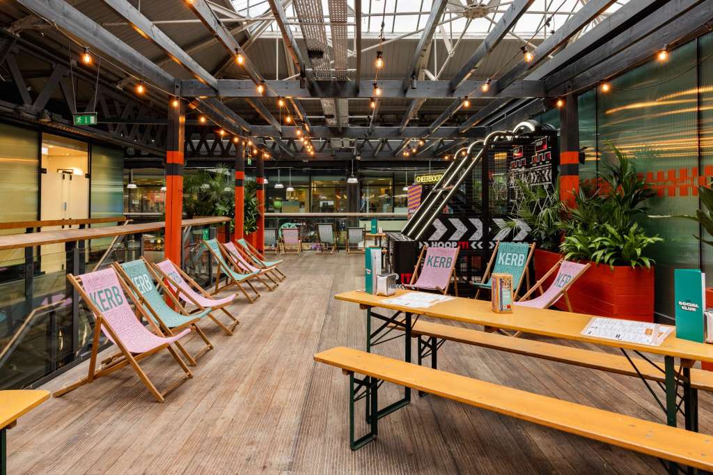 Modern sports bar interior with high ceilings, industrial beams, pendant lighting, branded deck chairs, and communal wooden tables.