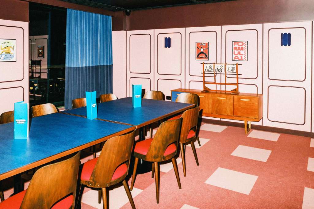 Modern meeting room with long blue table, wooden chairs with red seats, and pink paneled walls with artwork.