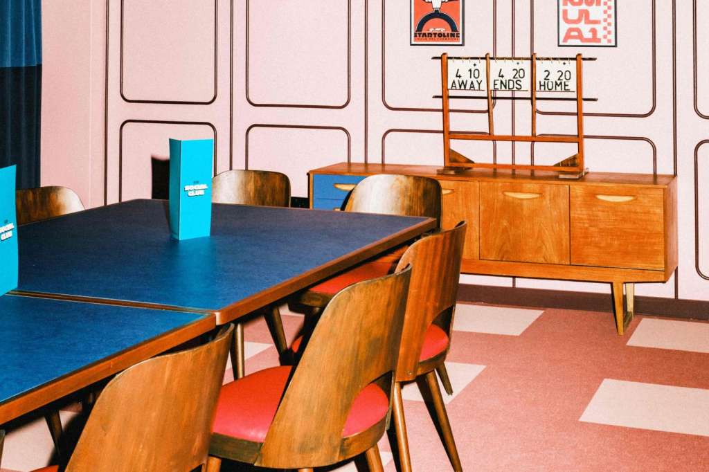 Interior space with blue table, wooden chairs, sports posters, and retro decor on pink and white flooring.