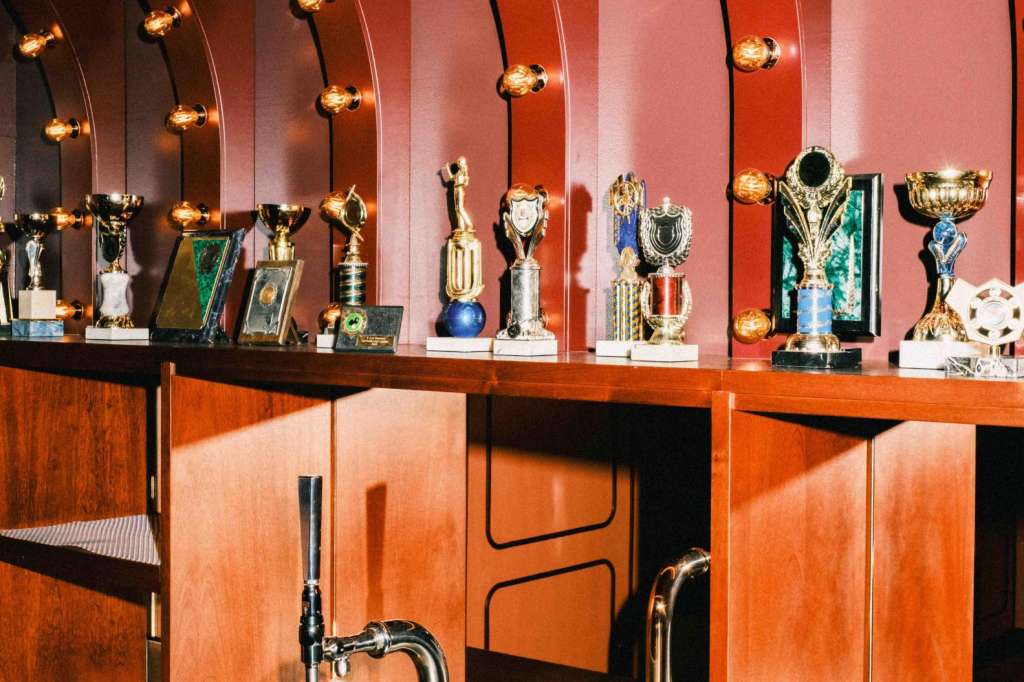 Bar counter with trophy display, warm Edison bulb lighting, and curved red-orange architectural backdrop.