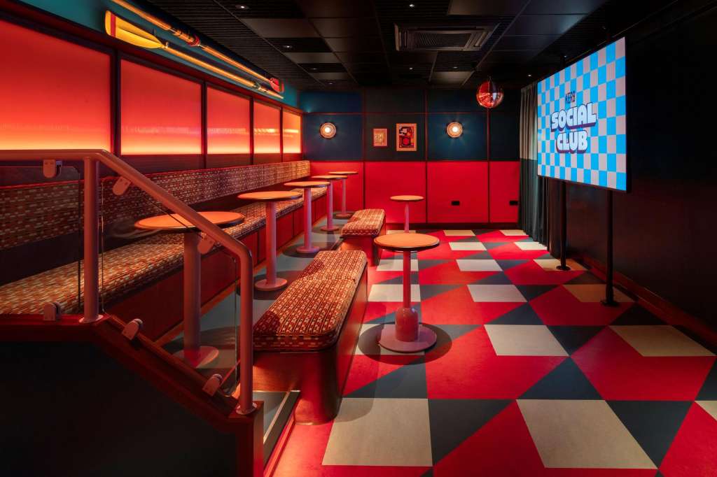 The Pitstop room with red and black geometric flooring, booth seating, high-top tables, and digital screen displaying social club branding.