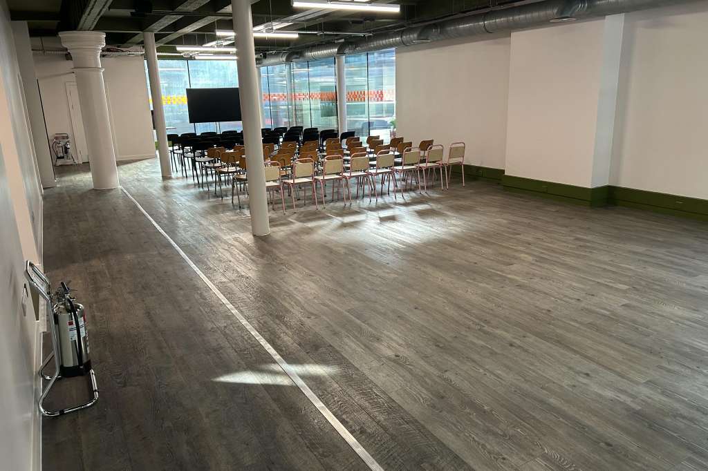 Large open event space with rows of chairs, support columns, and floor-to-ceiling windows overlooking a street.