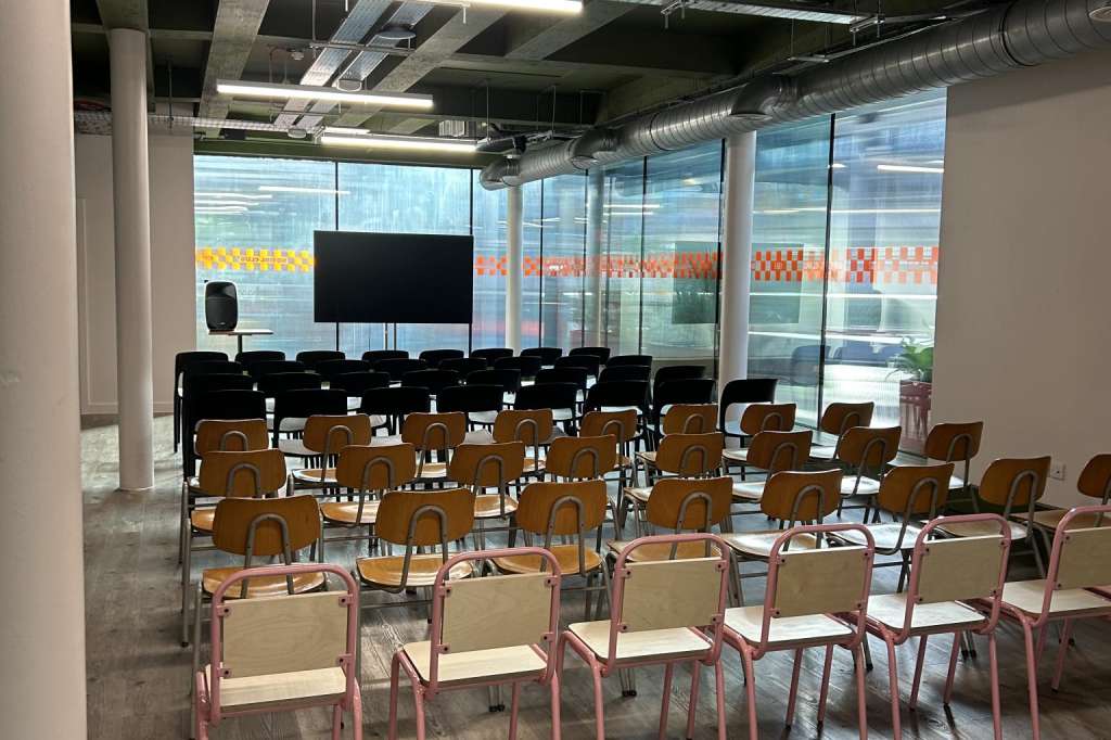 Modern conference space with rows of chairs facing a screen, exposed concrete ceiling, and floor-to-ceiling windows.