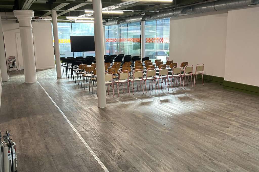 Modern event space with rows of chairs, exposed ceiling beams, support columns, and floor-to-ceiling windows overlooking a street.
