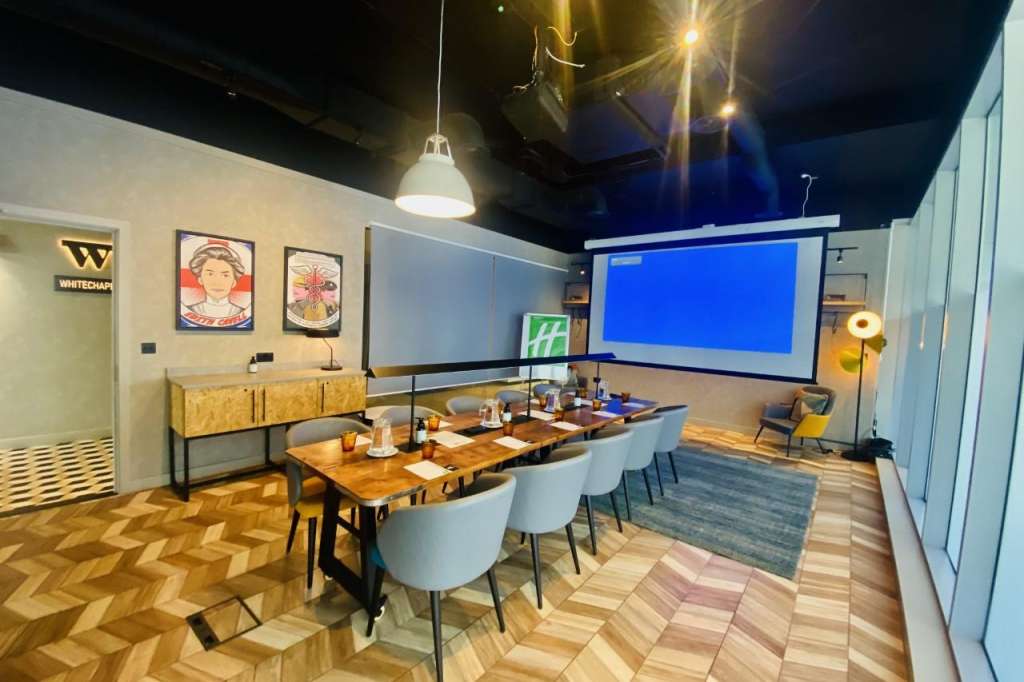 Modern meeting room with long wooden table, chairs, projection screen, pendant lighting, and contemporary artwork on walls.