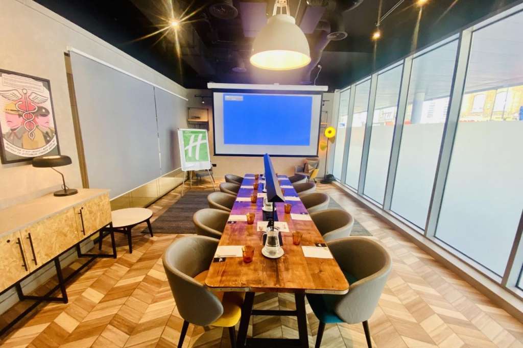 Modern meeting room with long wooden tables, grey chairs, projection screen, and pendant lighting.