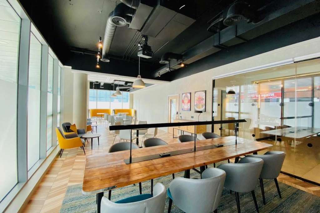 Modern meeting space with wooden tables, gray chairs, glass partitions, and industrial ceiling with pendant lighting.