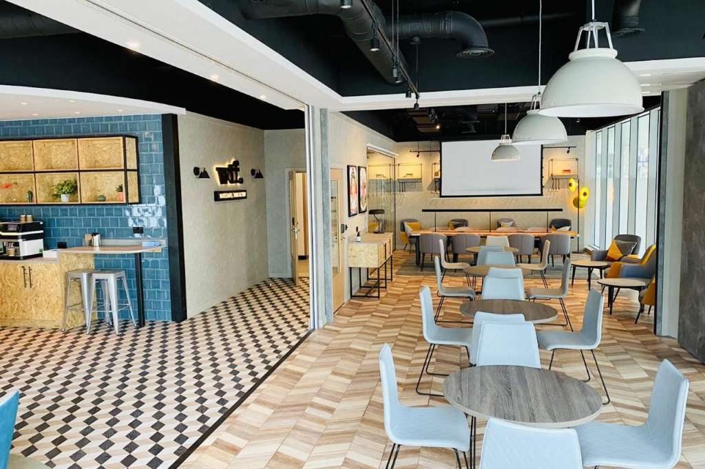 Modern open-plan meeting space with round tables, light blue chairs, kitchenette, and projection screen.