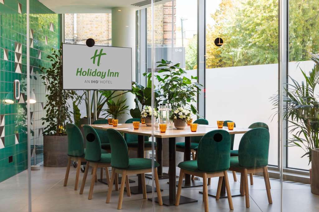 Modern meeting space with rectangular table, green chairs, potted plants, and floor-to-ceiling windows overlooking greenery.