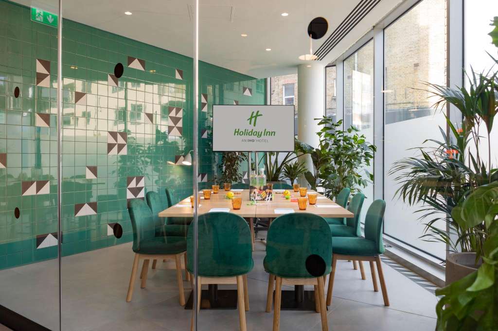 Modern meeting room with rectangular table, green chairs, geometric wall tiles, and potted plants by windows.