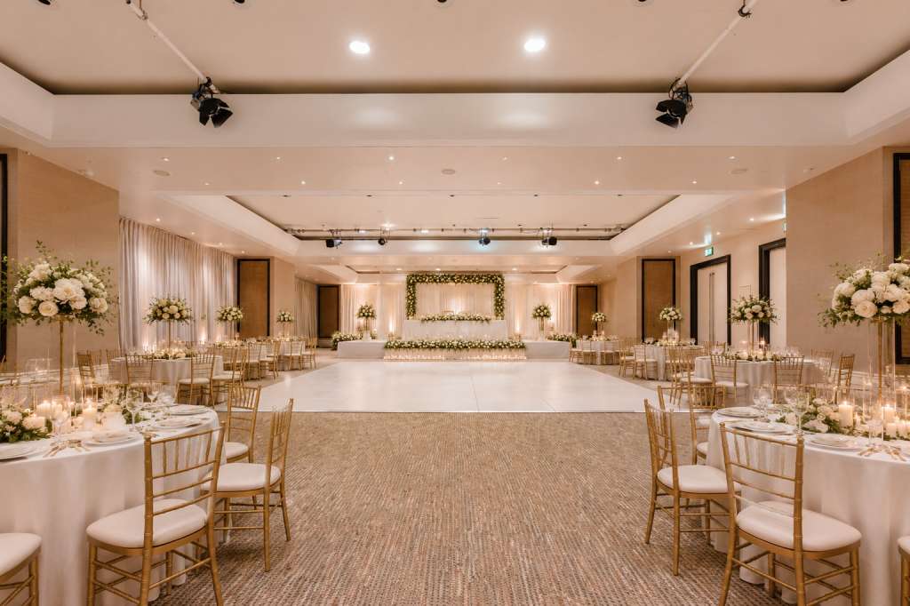 Ballroom set for a formal dinner with round tables, chiavari chairs, white linens, and floral centerpieces.