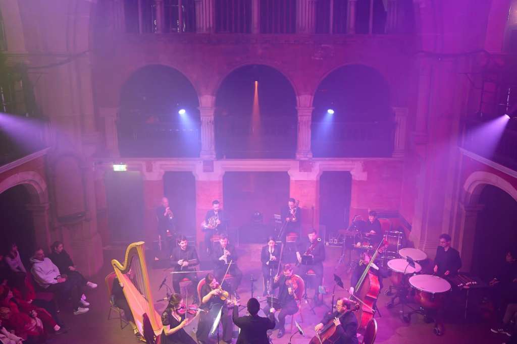 Grand hall with arched windows and organ, lit in purple, orchestra performing on stage with seated audience.