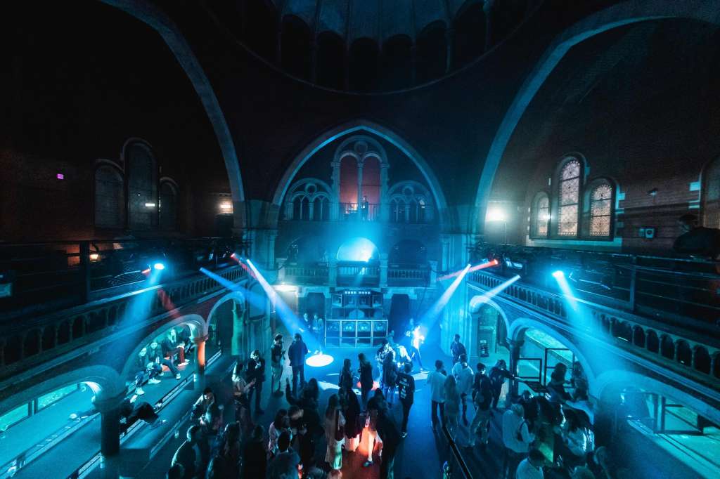 Grand historic interior with vaulted ceilings, dramatic blue lighting, and crowds gathered around illuminated installations.