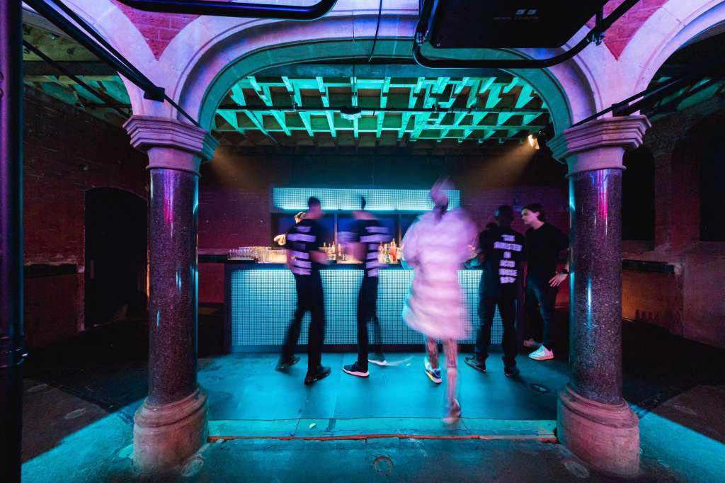 Modern venue with neon lighting, bar counter, and guests dancing under geometric ceiling panels.