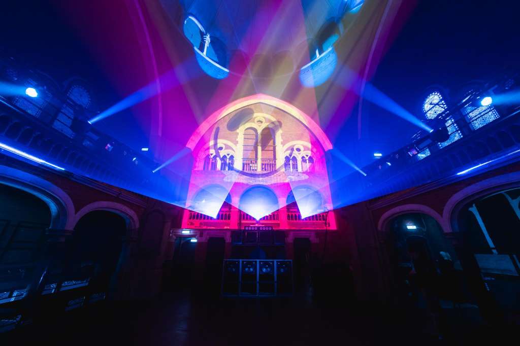 Grand interior space with ornate arched architecture illuminated by vibrant blue and pink stage lighting and projection.