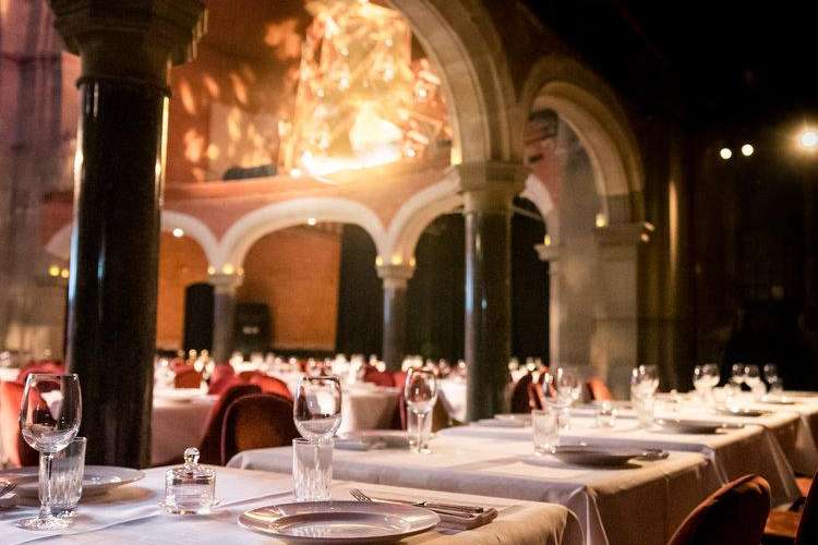 Elegant outdoor courtyard set for a formal dinner with long tables, white linens, place settings, and arched stone architecture illuminated at night.