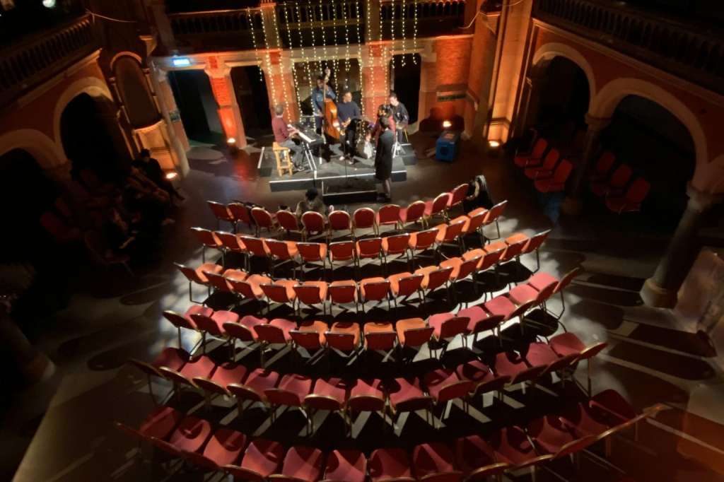 Grand auditorium with curved rows of red seats facing a lit stage with architectural arches and decorative lighting.