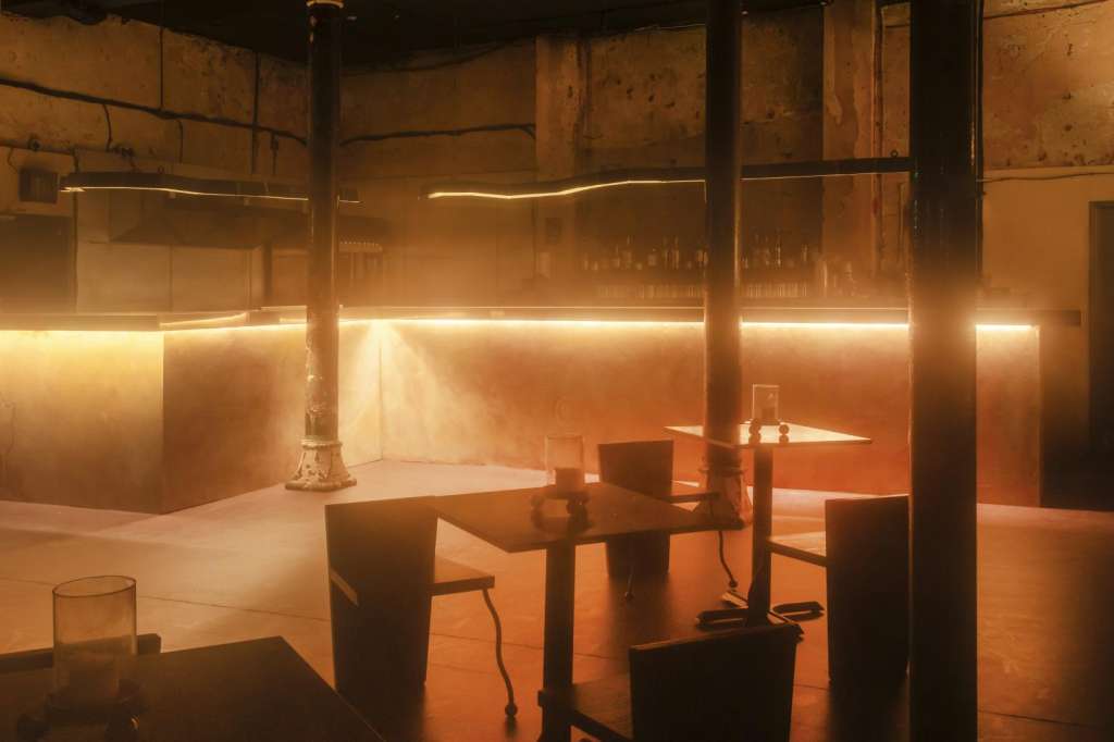 Industrial space with warm ambient lighting, scattered tables and chairs, exposed brick walls and steel columns.