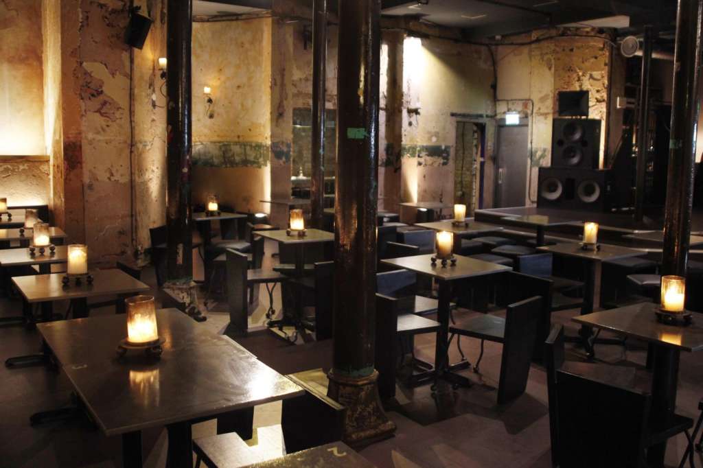 Industrial venue with exposed brick walls, black tables, candlelit ambiance, and stage speakers.